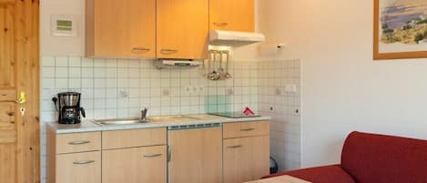 Cocina compacta con armarios de madera, cafetera y lavamanos. En la zona de estar contigua, hay un sofá rojo.