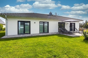 Ein modernes, zweistöckiges Haus mit flachem Dach, großen Fenstern und einer Terrasse mit Gartenmöbeln.