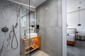 Een moderne badkamer met een douchecabine van glas, een houten wastafelmeubel en een zichtbare slaapkamer met een bed en een hanglamp.