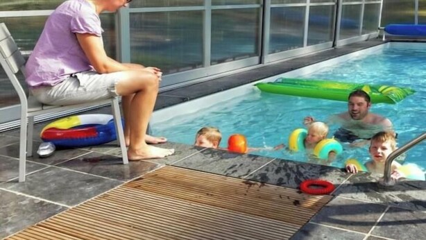 Une piscine entourée de parois en verre, une personne assise sur une chaise et des enfants qui jouent dans l’eau.