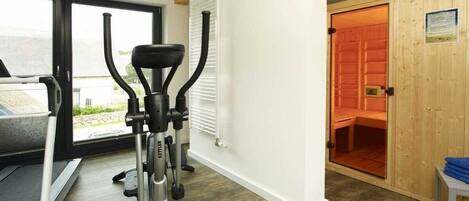 Ein Fitnessraum mit Laufband, Crosstrainer und einer Sauna, die durch eine Tür sichtbar ist.
