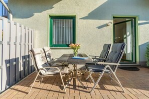 Une terrasse extérieure avec une table et deux chaises, un petit vase avec des fleurs et un bâtiment doté d’une fenêtre et d’une porte vertes.