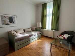 Ein Zimmer mit einem Bett, einem Sessel, einem Tisch, einer Lampe und einem Fenster mit Vorhängen.