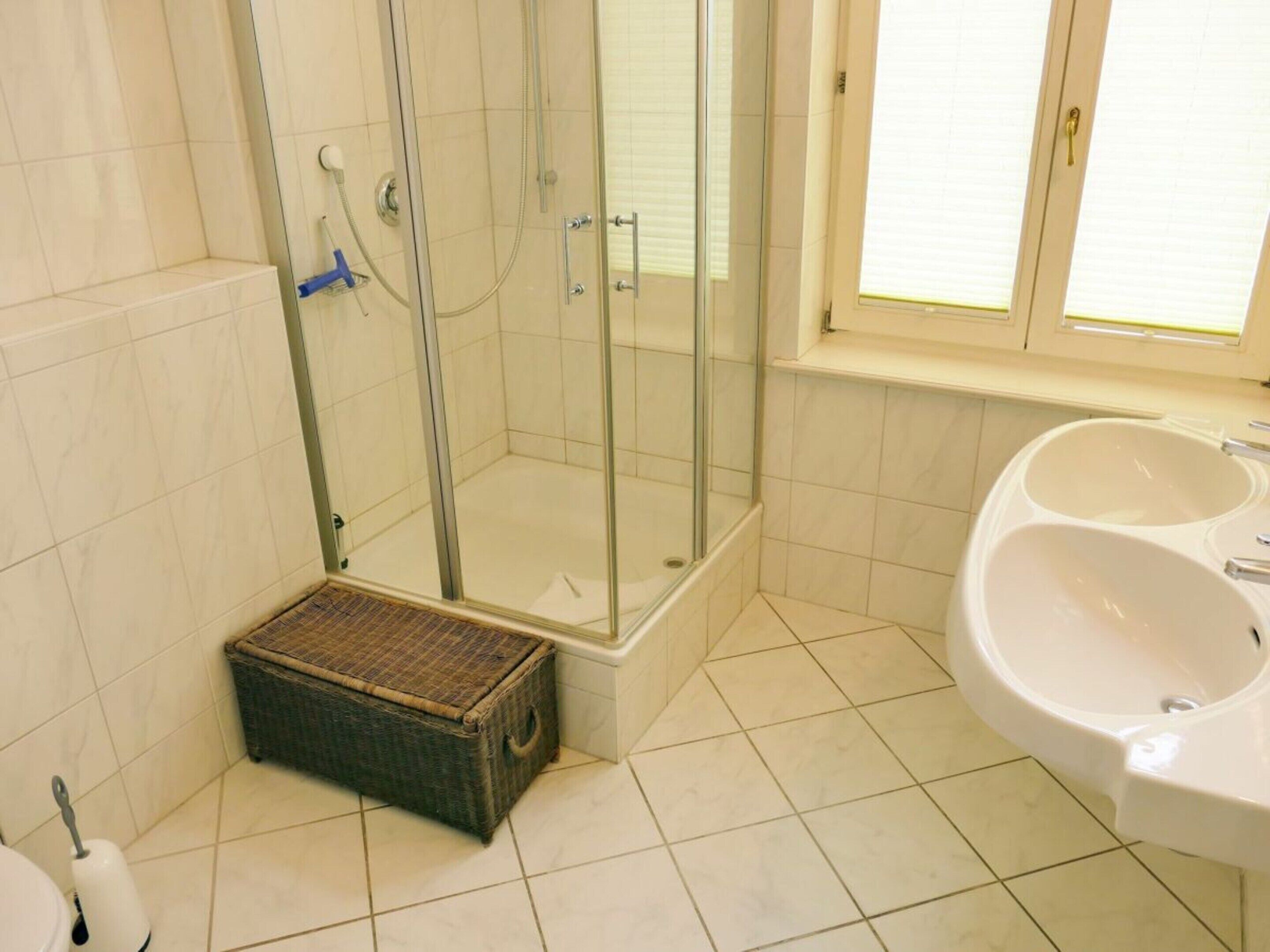 A bathroom with a glass-enclosed shower, a white sink with dual basins, and a wicker basket on the floor.