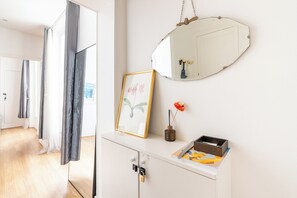 Ein Flur mit einer weißen Tür, einem Spiegel, einem gerahmten Bild, einer Vase mit einer einzelnen Blume und einer kleinen Holzschatulle auf einem weißen Schrank.