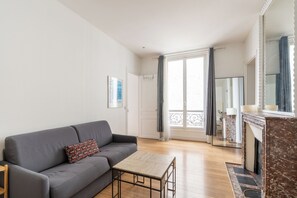 A living room with a grey sofa, a small wooden table, a fireplace, and a large window with curtains. - Cozy 1BDR Apartment for 4 Near Champs-Elysees (Paris)