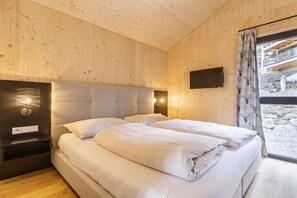 Ein Schlafzimmer mit Holzboden, einem Bett mit weißer Bettwäsche, einem Nachttisch, einem an der Wand montierten Fernseher und einem Fenster mit Vorhängen.