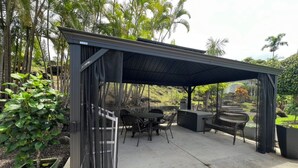 Área de estar coberta ao ar livre, com mesa e cadeiras, cercada por vegetação e palmeiras.