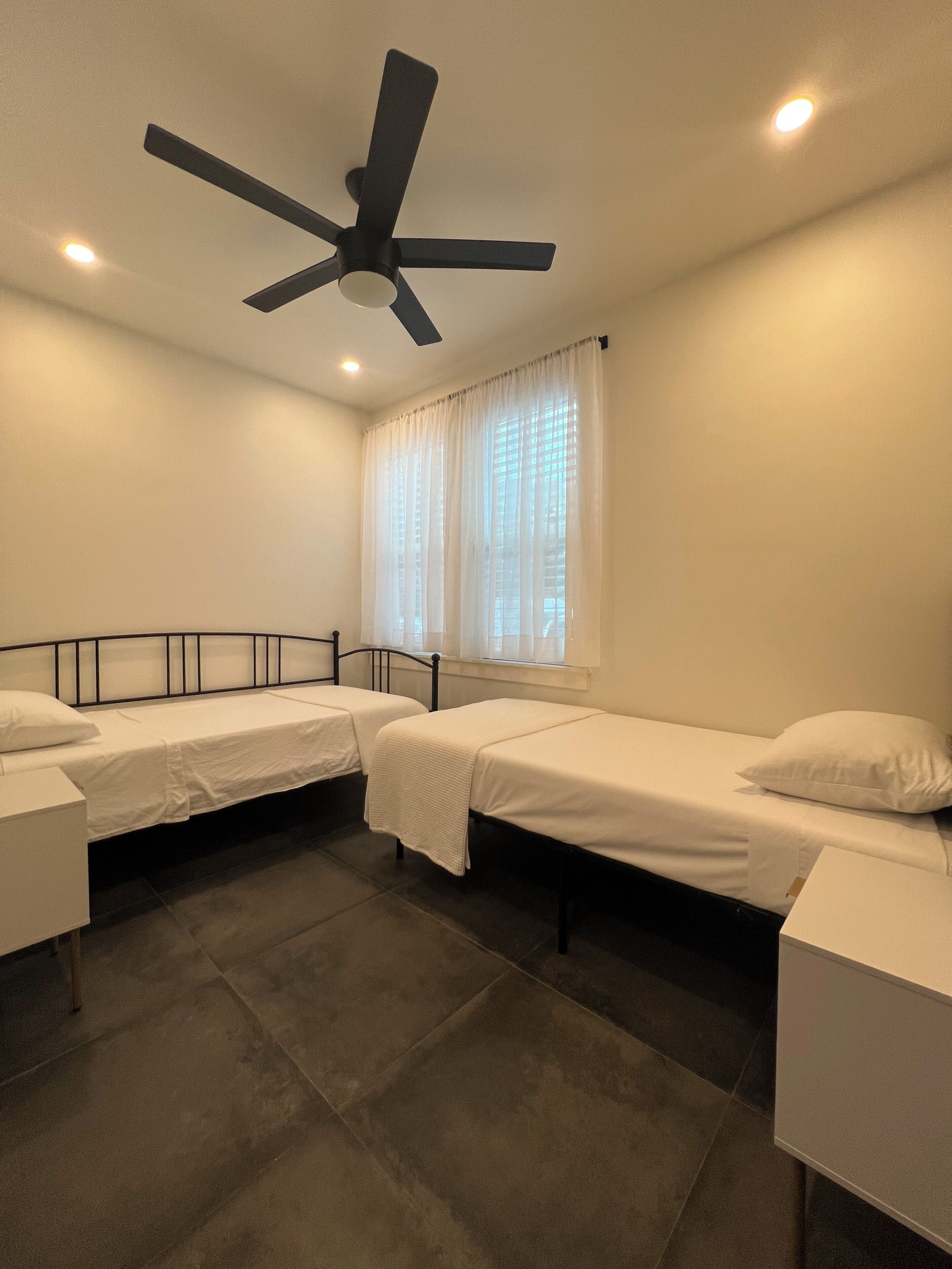 A room with two single beds, a ceiling fan, a window with curtains, and a tiled floor.