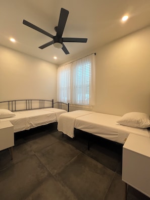 Une chambre avec deux lits simples, un ventilateur de plafond, une fenêtre avec des rideaux et un sol carrelé.