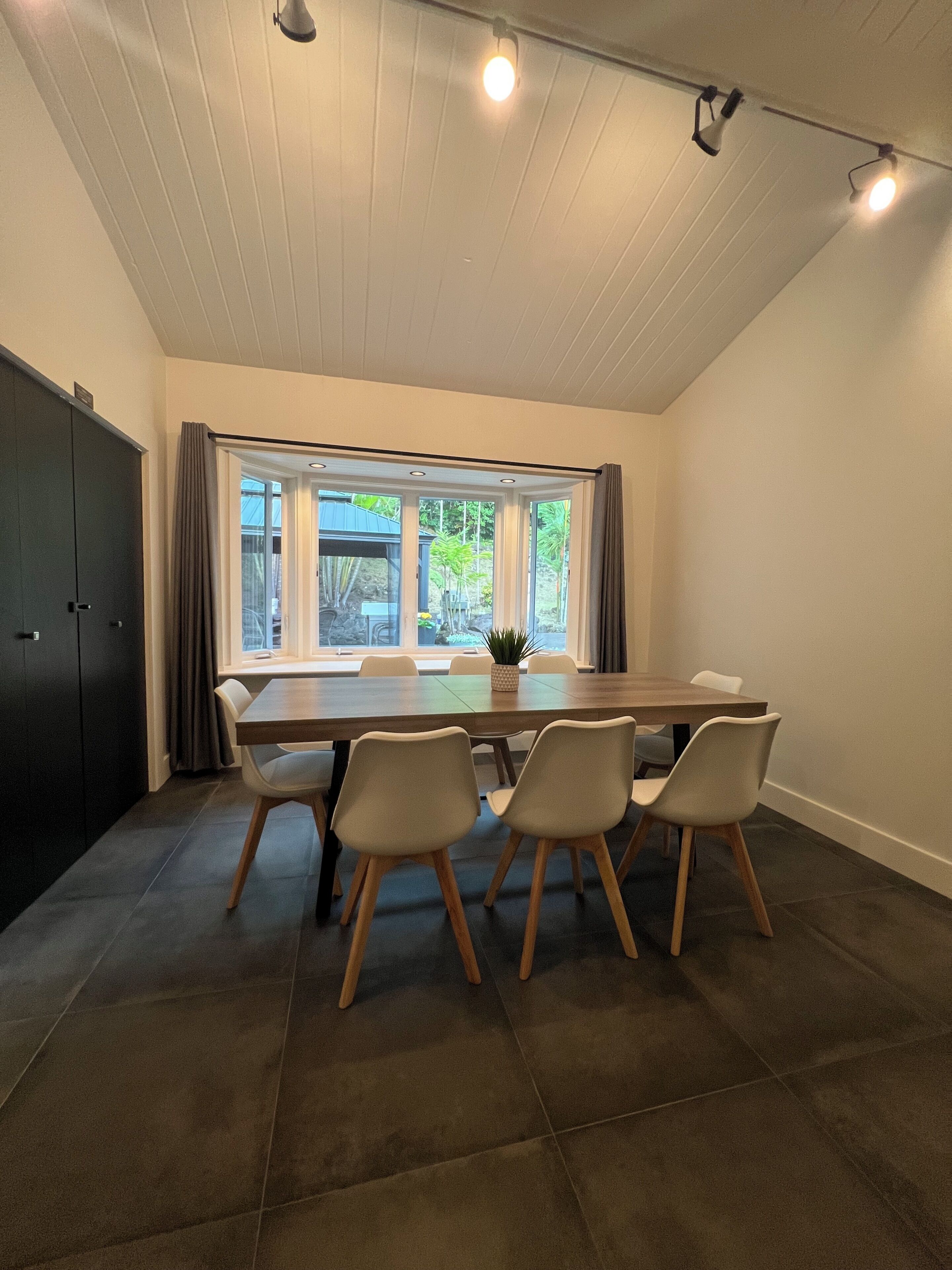 A modern dining area with a long table, four chairs, and a view of the outdoors through large windows.