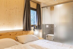 Une chambre à coucher avec une tête de lit en bois, un lit avec une literie blanche, une fenêtre avec des rideaux et une salle de bain équipée de toilettes et d’un porte-serviettes.