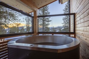 A wooden cabin with a large, round hot tub in the center, surrounded by large windows offering a view of trees and mountains. - Superior Chalet # 8 with Sauna & Hot Tub (Turrach)