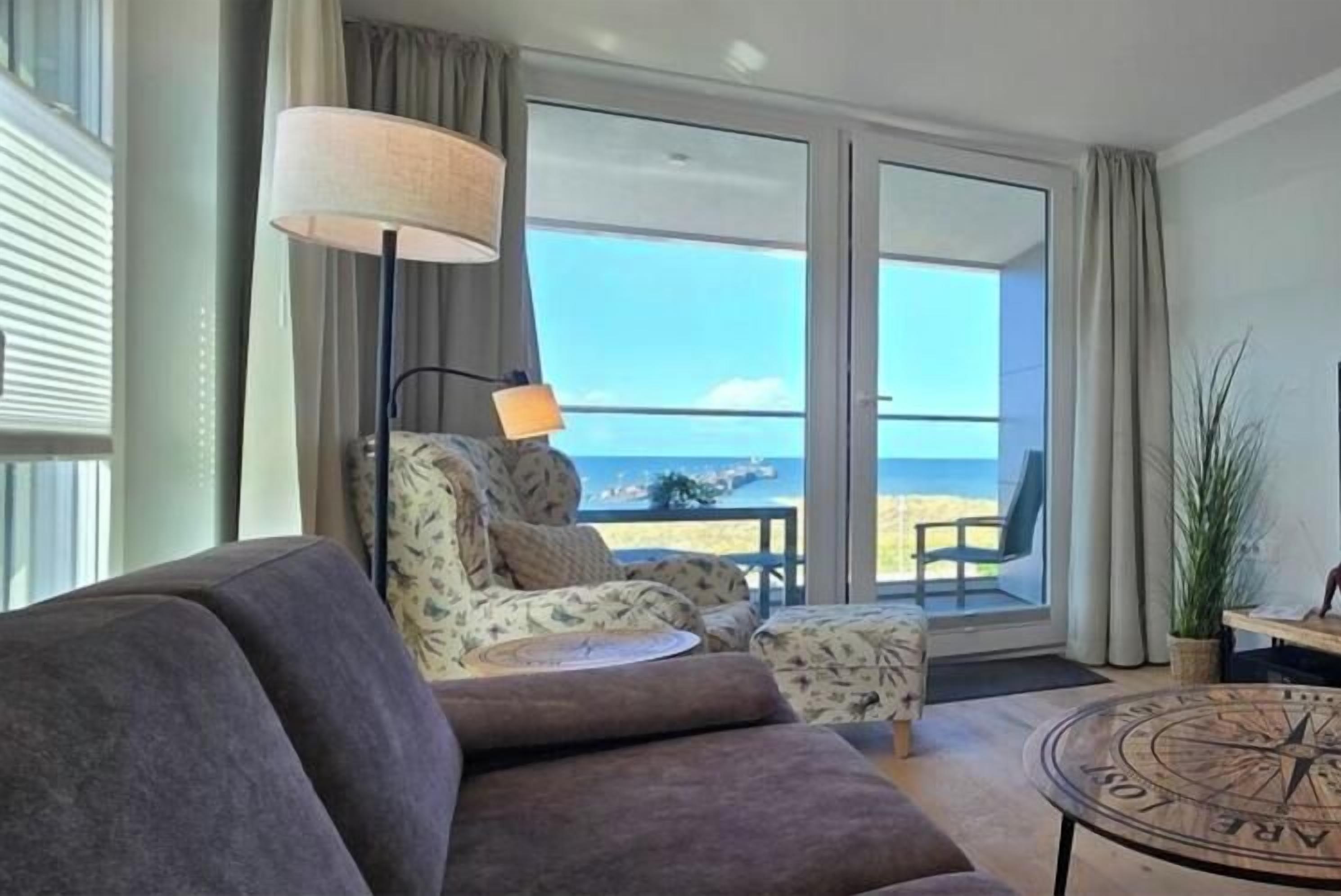 Uma sala de estar com sofá, poltrona e mesa de centro. Há uma janela grande com vista para a praia.
