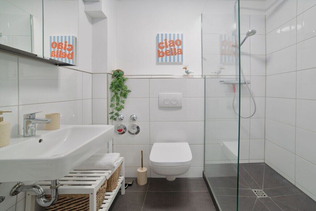 Un baño moderno con lavamanos, inodoro y ducha blancos. Hay dos toalleros y una planta pequeña en la pared.