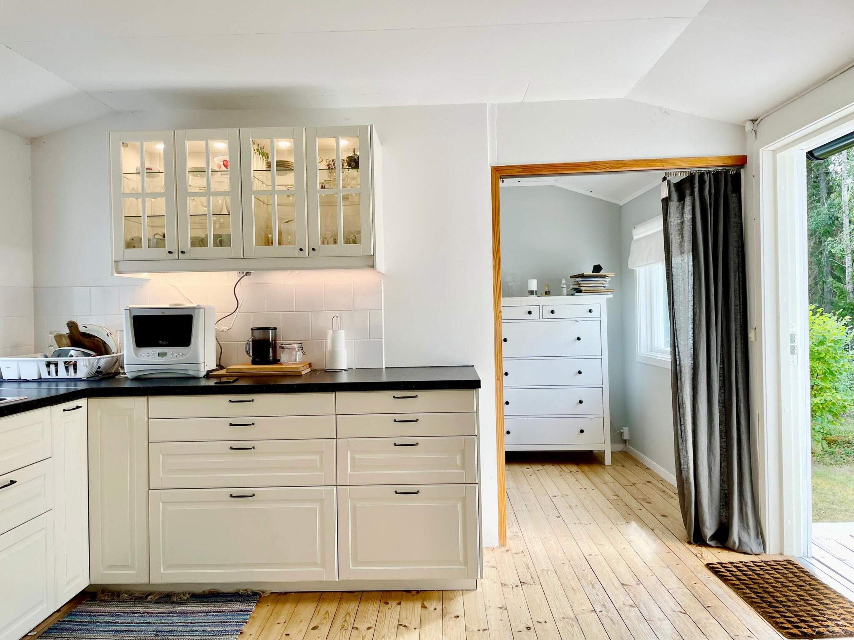 A kitchen with white cabinets, a microwave, and a coffee maker. There is a window with curtains and a view of greenery outside.