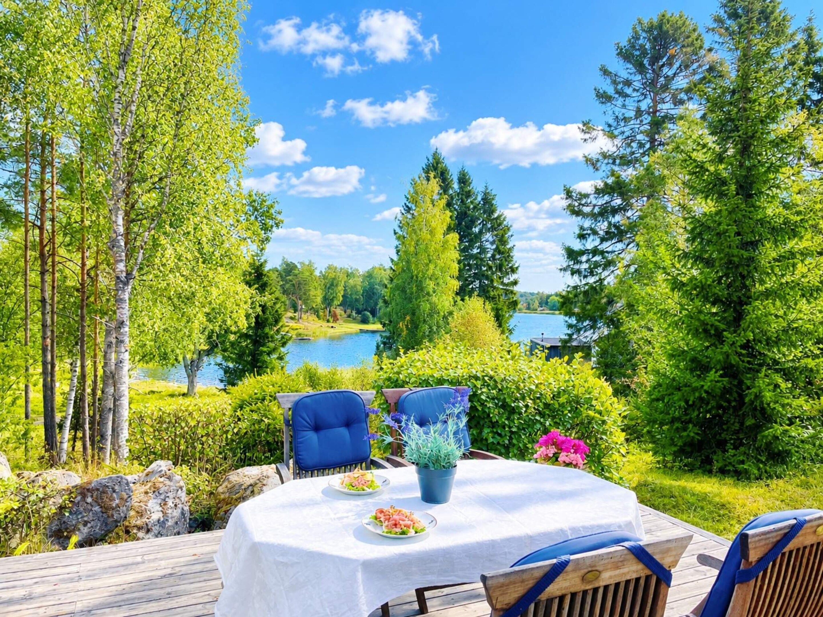 An outdoor dining area with a table and chairs, surrounded by greenery and a view of a lake.