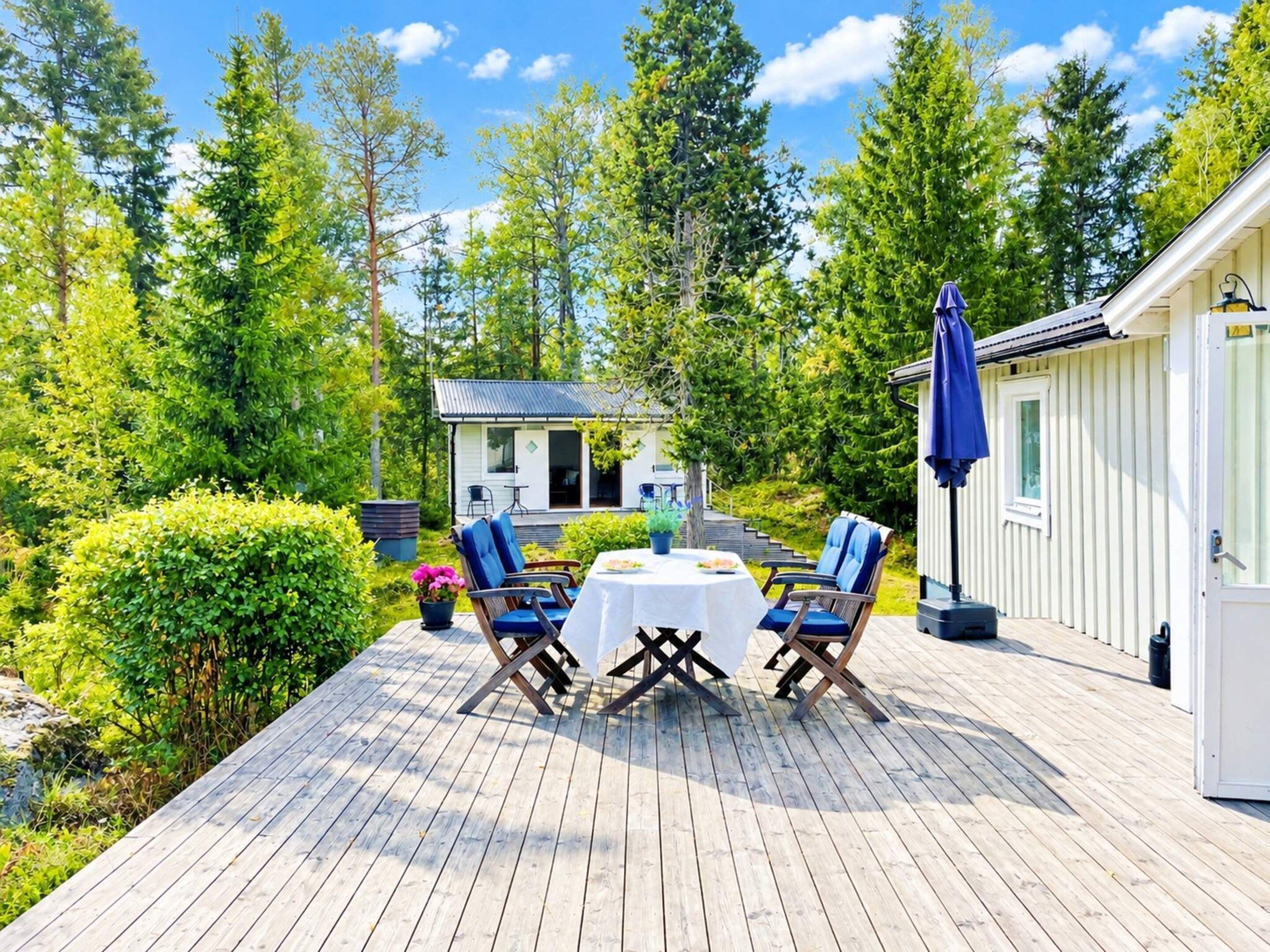 Eine Holzterrasse mit Tisch und Stühlen, umgeben von Grünpflanzen und einem kleinen Gebäude.