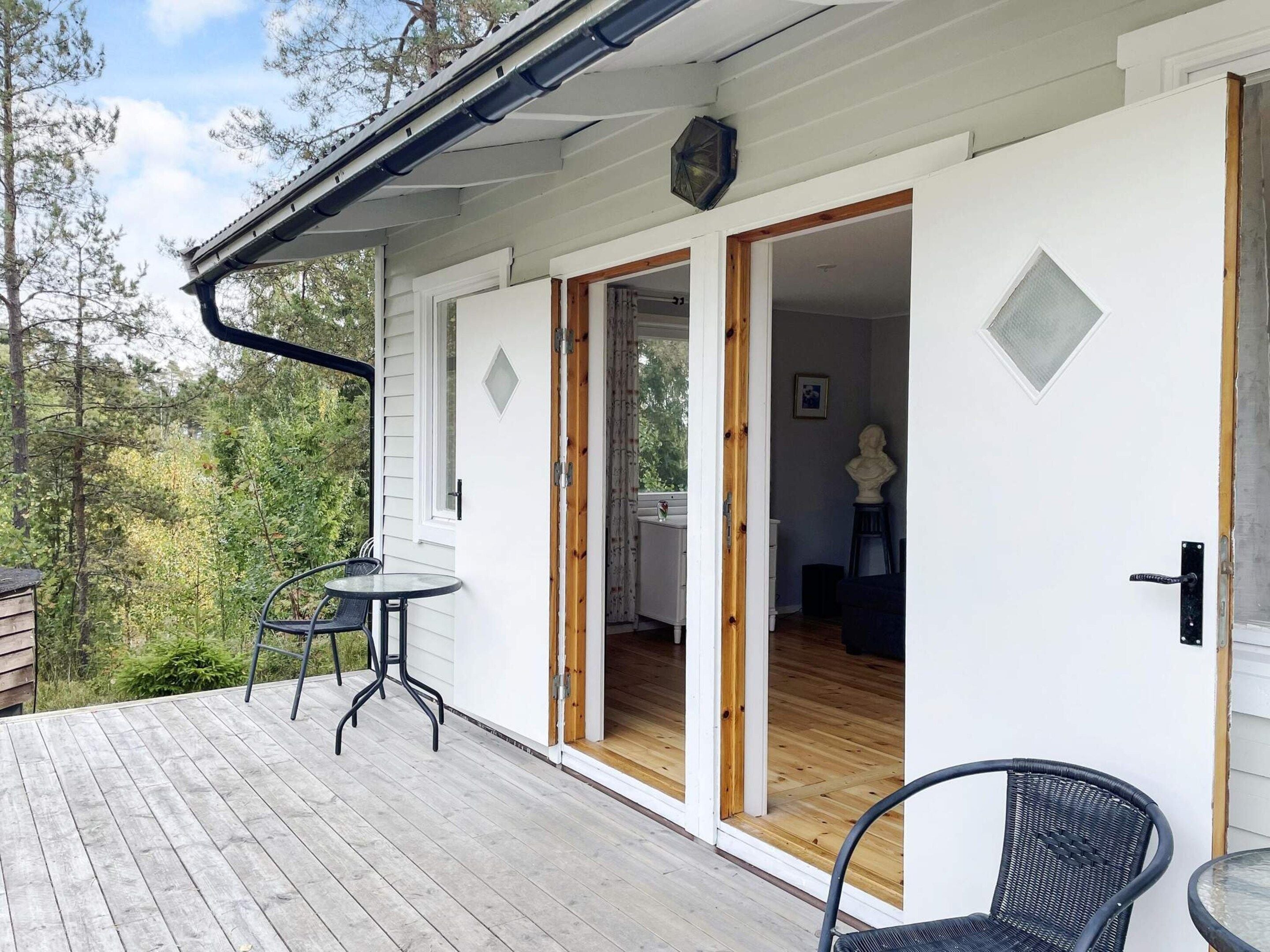 A wooden deck with a table and chairs, a white exterior wall, and a door leading to an interior space.