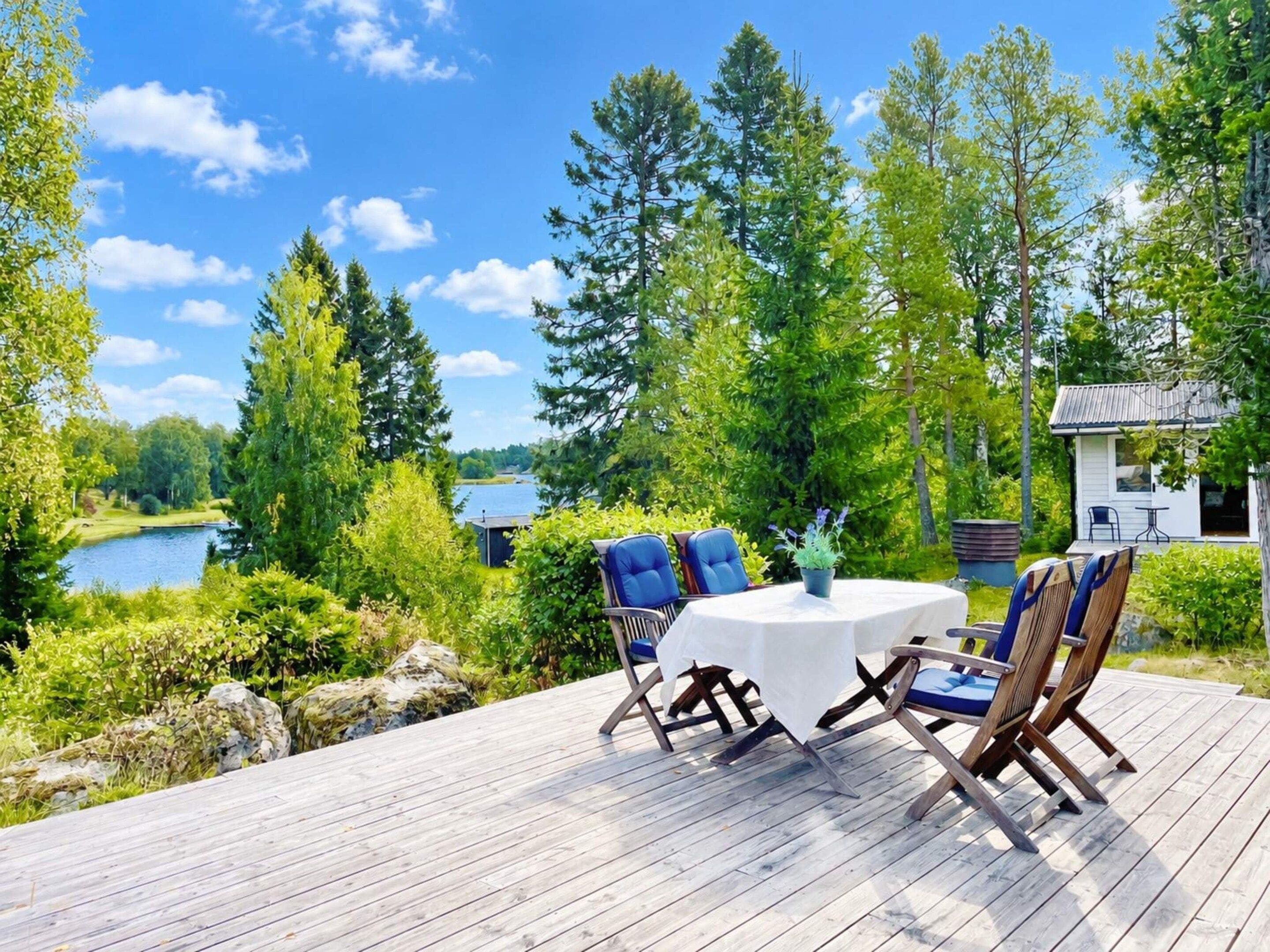 Eine Terrasse mit Gartenmöbeln, die auf einen See mit umliegenden Bäumen und einem kleinen Gebäude blickt.