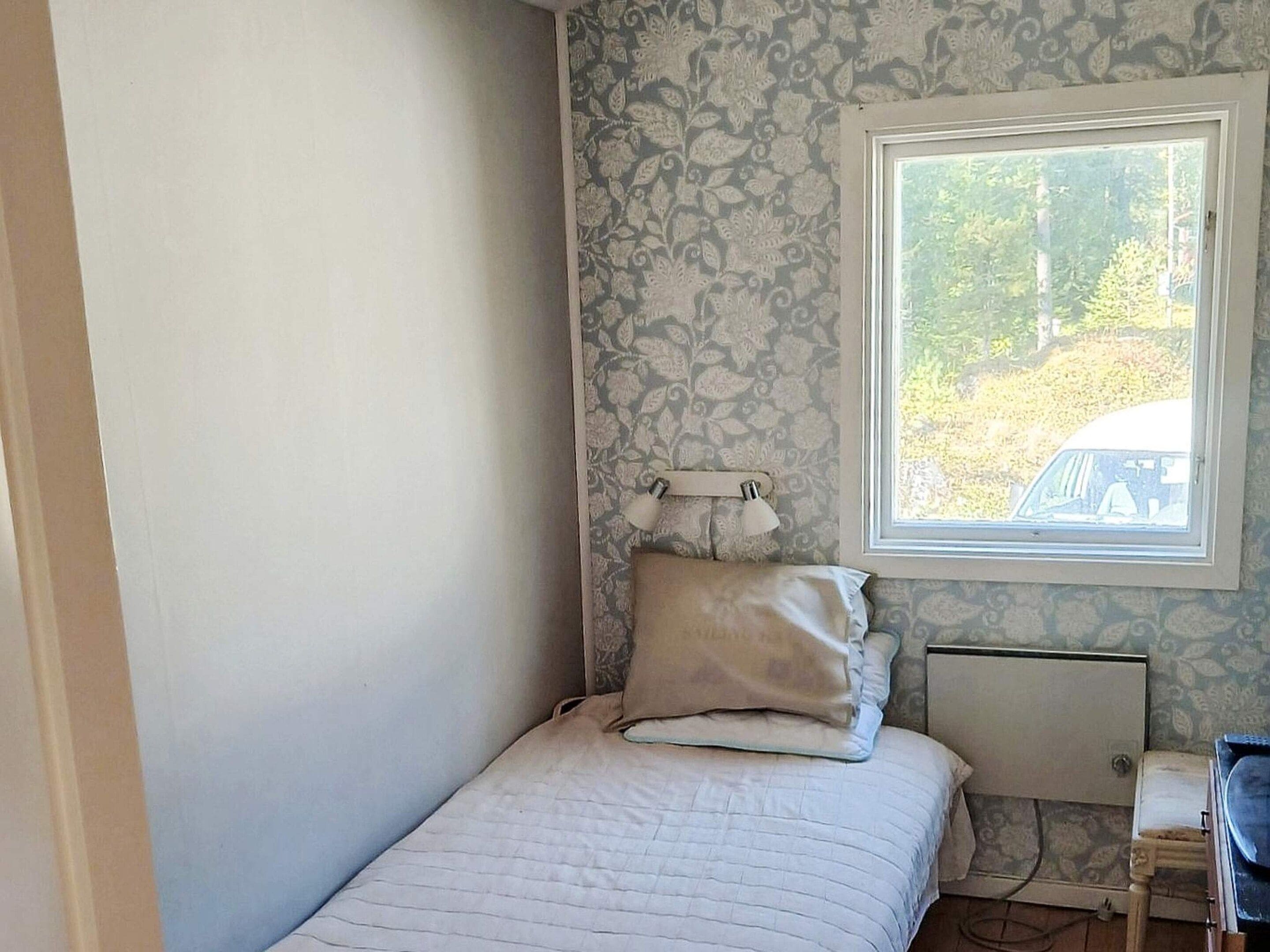 Un dormitorio pequeño con una cama individual, una mesita de noche, una lámpara y una ventana que da a vistas de árboles y un vehículo fuera.