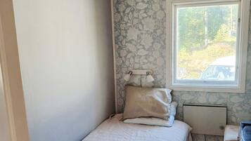 Un dormitorio pequeño con una cama individual, una mesita de noche, una lámpara y una ventana que da a vistas de árboles y un vehículo fuera.