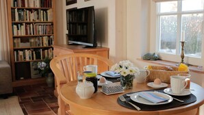 A dining area with a round wooden table set for breakfast, a bookshelf, a television, and a window with a view of trees. - Holiday home with 1 bedroom (Ostseebad Wustrow)