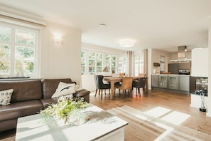 A modern living room with a brown leather sofa, a dining table with chairs, and a kitchen area with cabinets and a stove. - Holiday home with 3 bedrooms (Ostseebad Prerow)