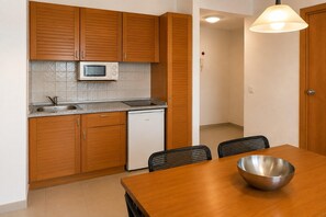 Een compacte keuken met houten kastjes, een magnetron, een gootsteen en een vaatwasser. Er staat een eettafel met stoelen en een metalen kom op.