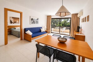 Een moderne woonkamer met een blauwe bank, een houten eettafel en stoelen. Er is een groot raam met gordijnen, een plafondlamp en een kleine tafel met een schaal.