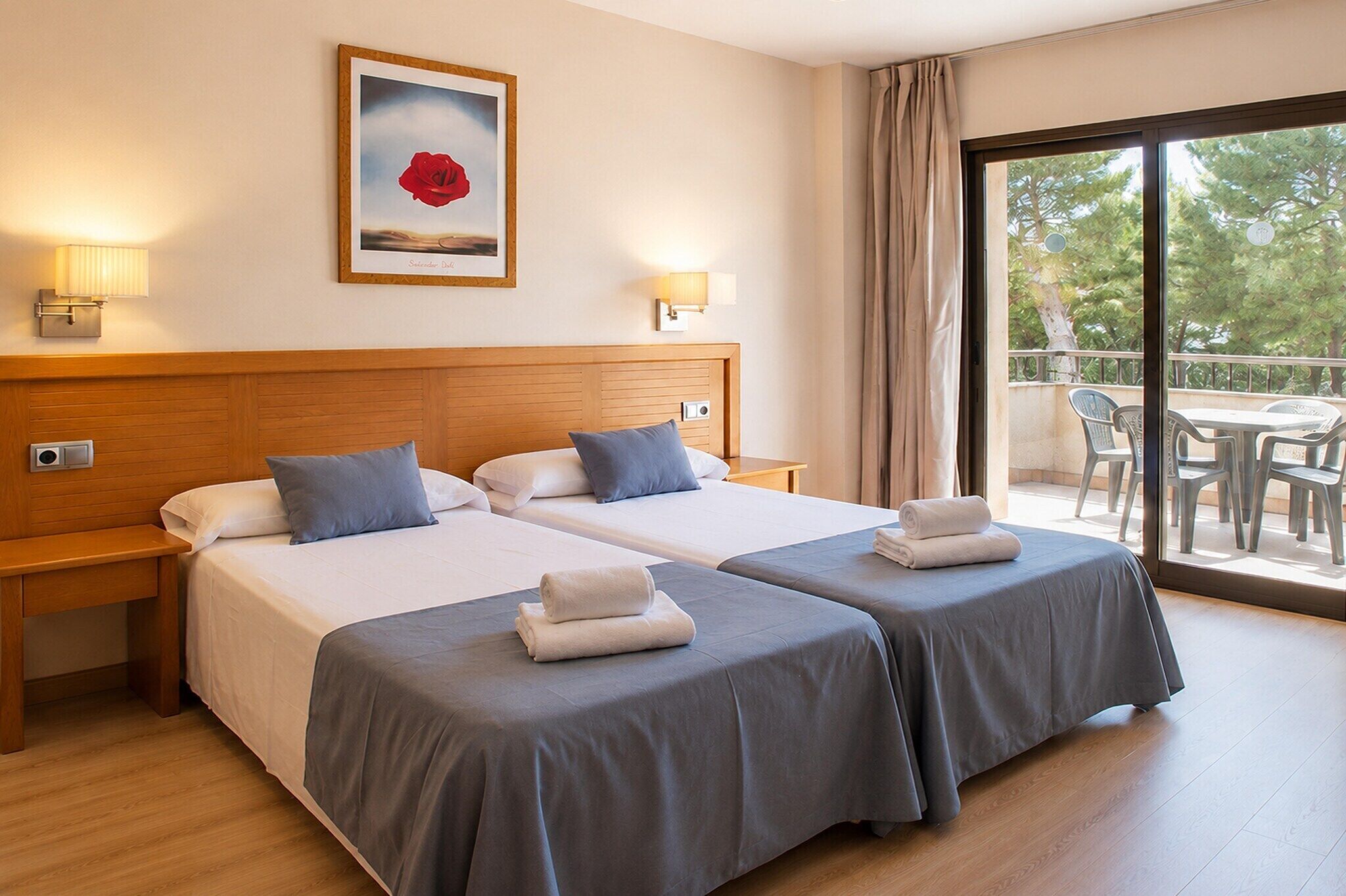 A hotel room with two beds, a wooden headboard, bedside lamps, a painting, and a balcony with chairs.