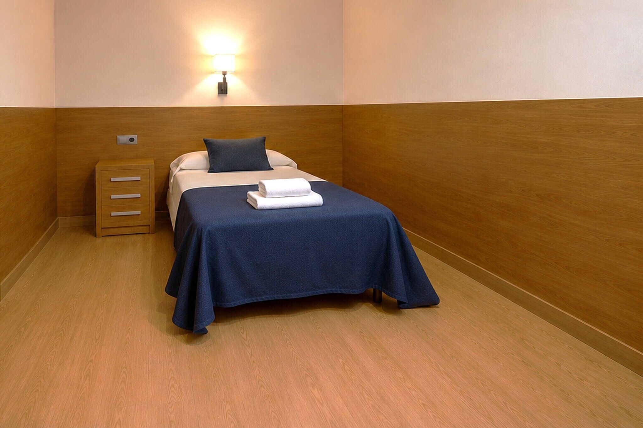 Een enkel bed met een blauwe sprei, een nachtkastje van hout met lades, en een wandlamp.
