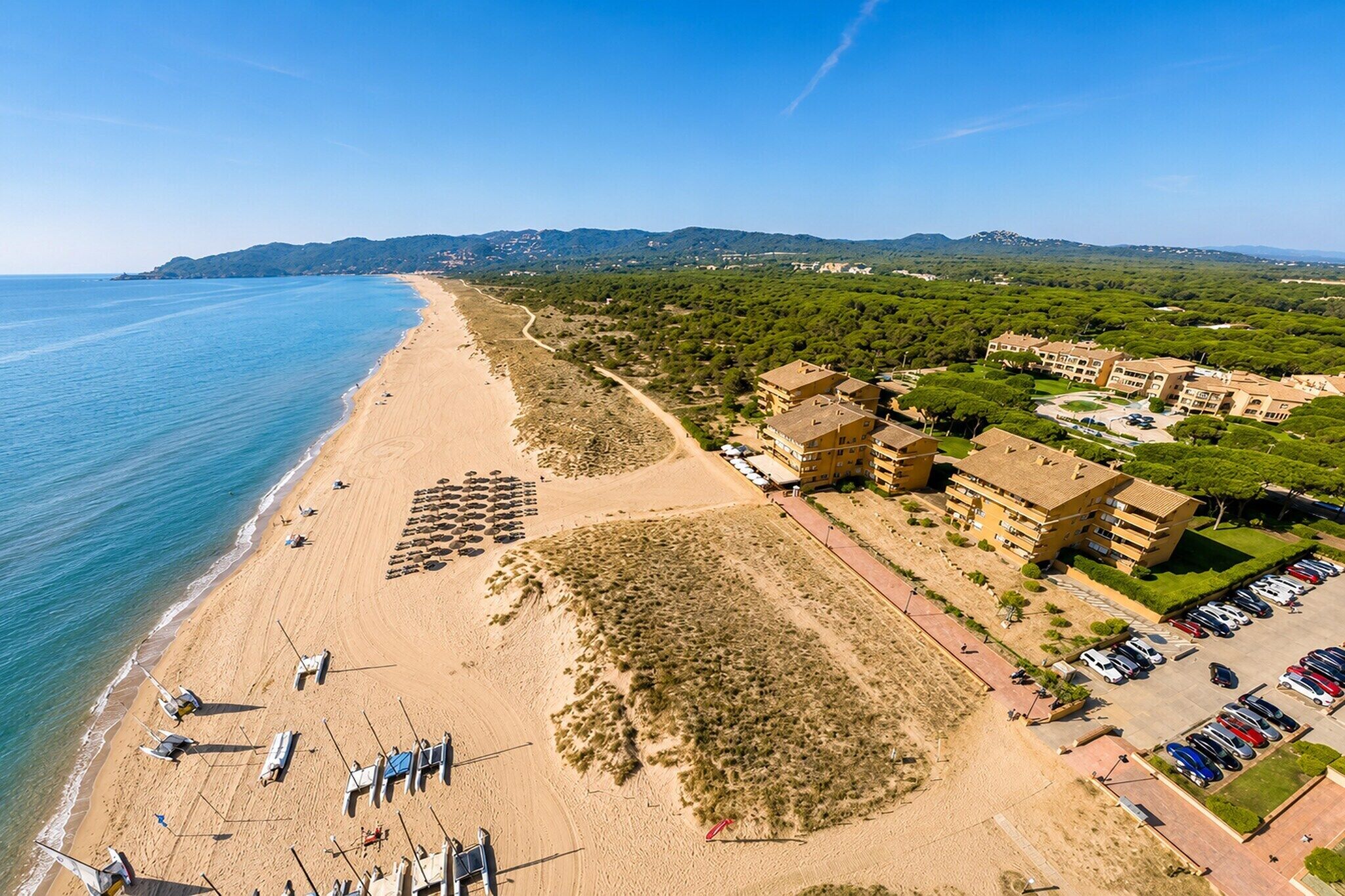 An aerial view of a coastal hotel with a beach, parking, and surrounding greenery.