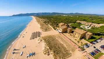 An aerial view of a coastal hotel with a beach, parking, and surrounding greenery.