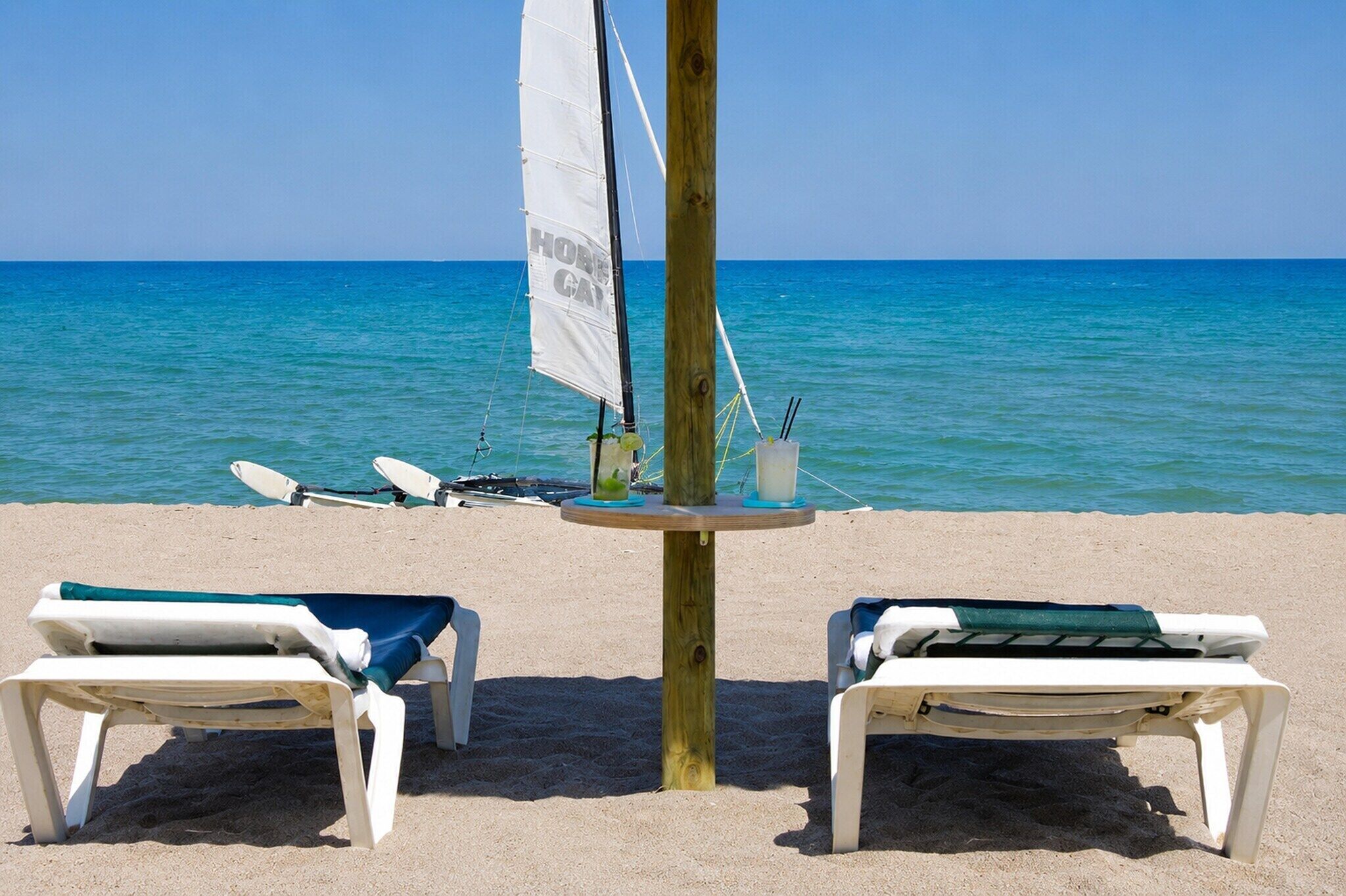 A beach with two lounge chairs, a table with a sail, and a surfboard.