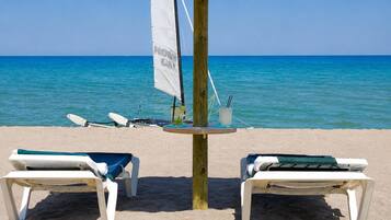 A beach with two lounge chairs, a table with a sail, and a surfboard.