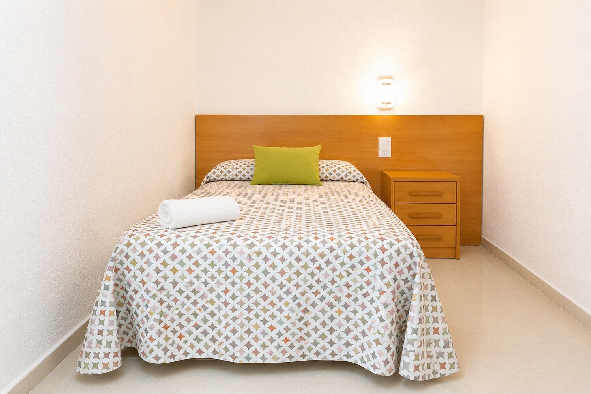 Een opgemaakt bed met een bedsprei met dessin, een groen kussen en een witte handdoek.
