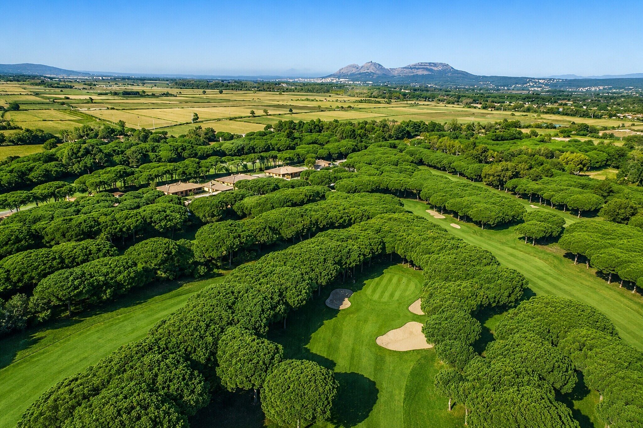 Luchtbeeld van een golfbaan met keurig bijgesnoeide bomen, zandbunkers en een bergketen in de verte.