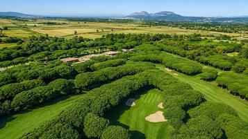 Luchtbeeld van een golfbaan met keurig bijgesnoeide bomen, zandbunkers en een bergketen in de verte.