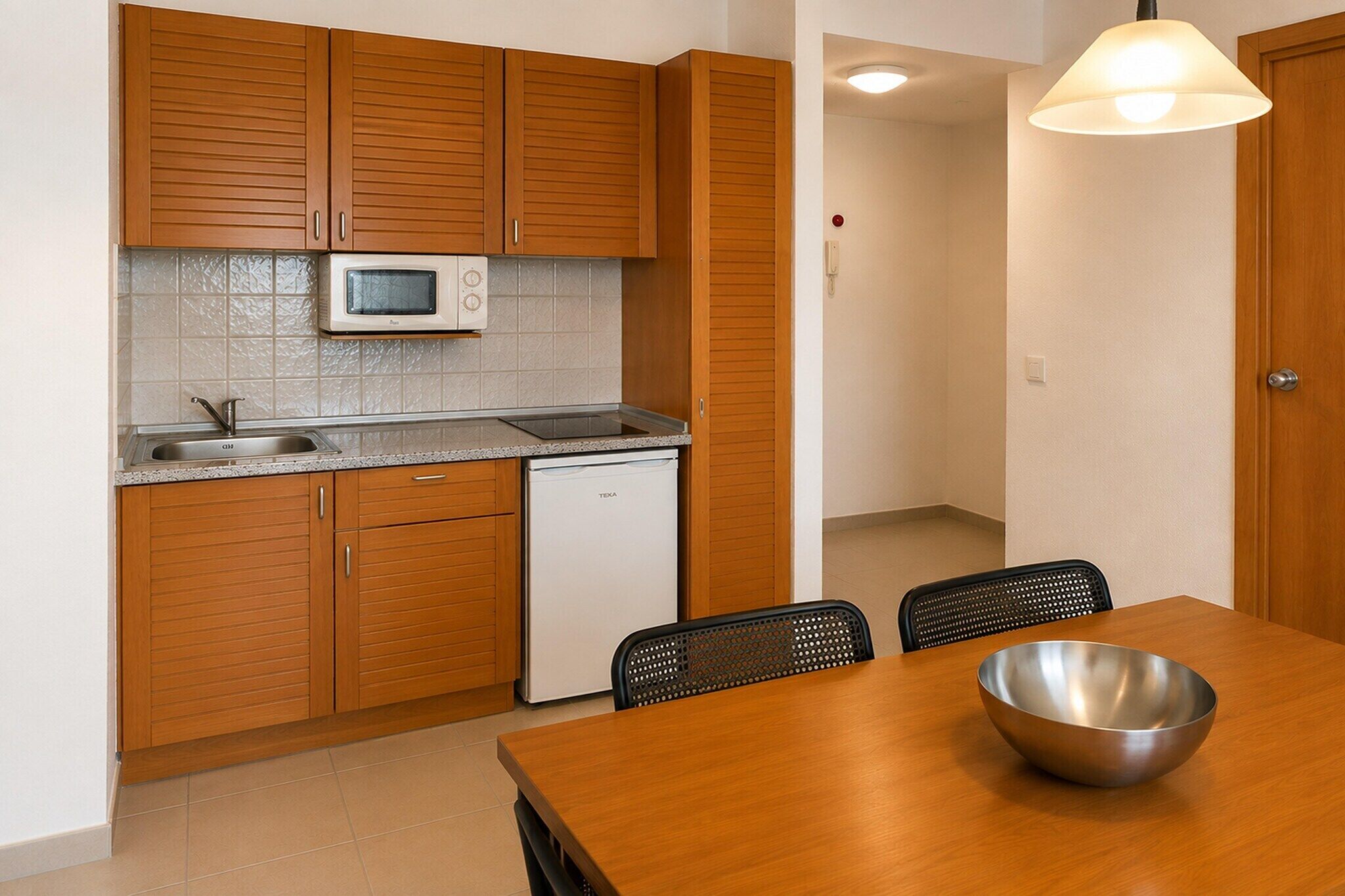 A compact kitchen with wooden cabinets, a microwave, a sink, and a dishwasher. There is a dining table with chairs and a metal bowl on it.