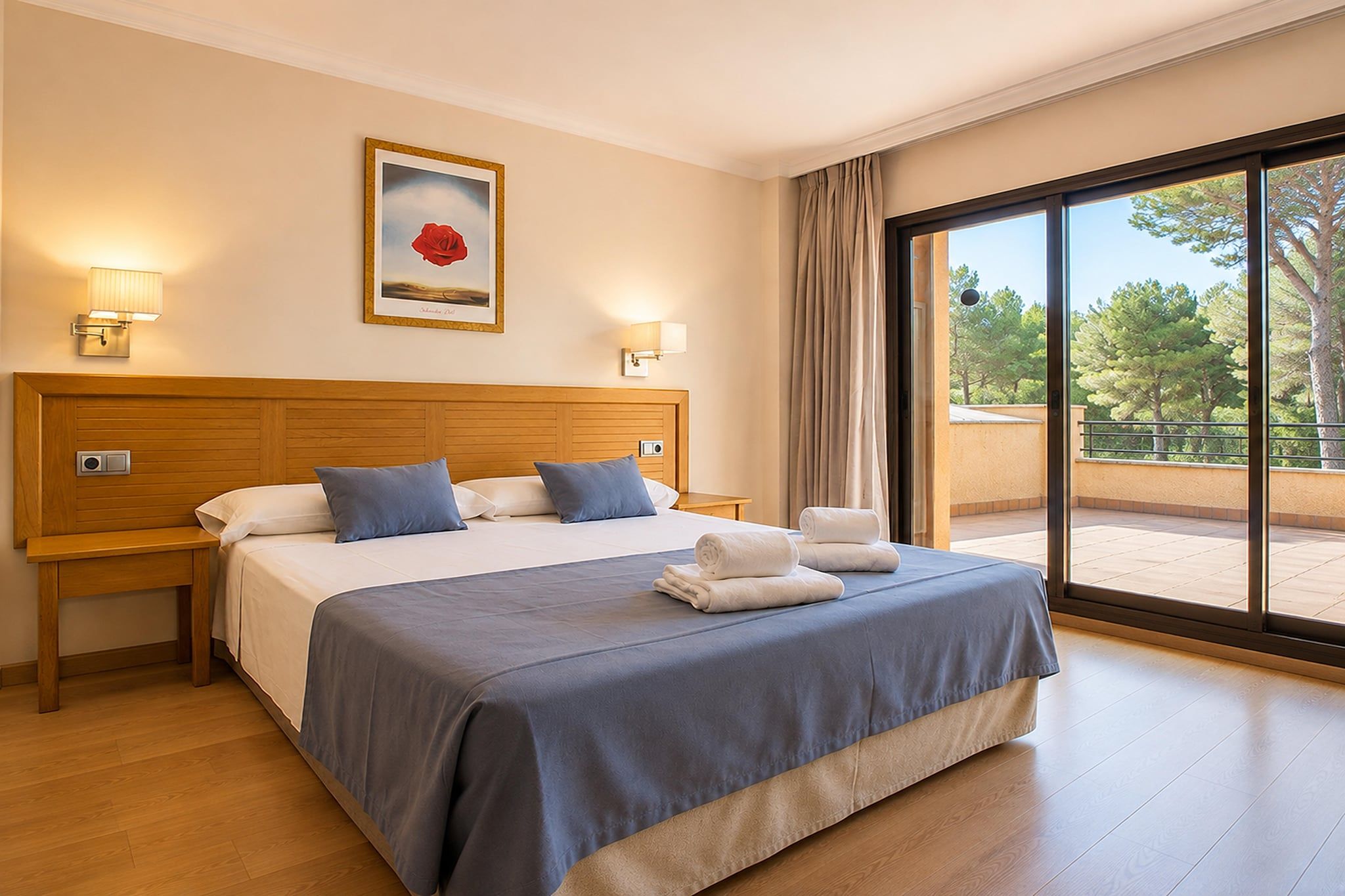 A hotel room with a large bed, two bedside tables, a painting, and a view of trees through a large window.