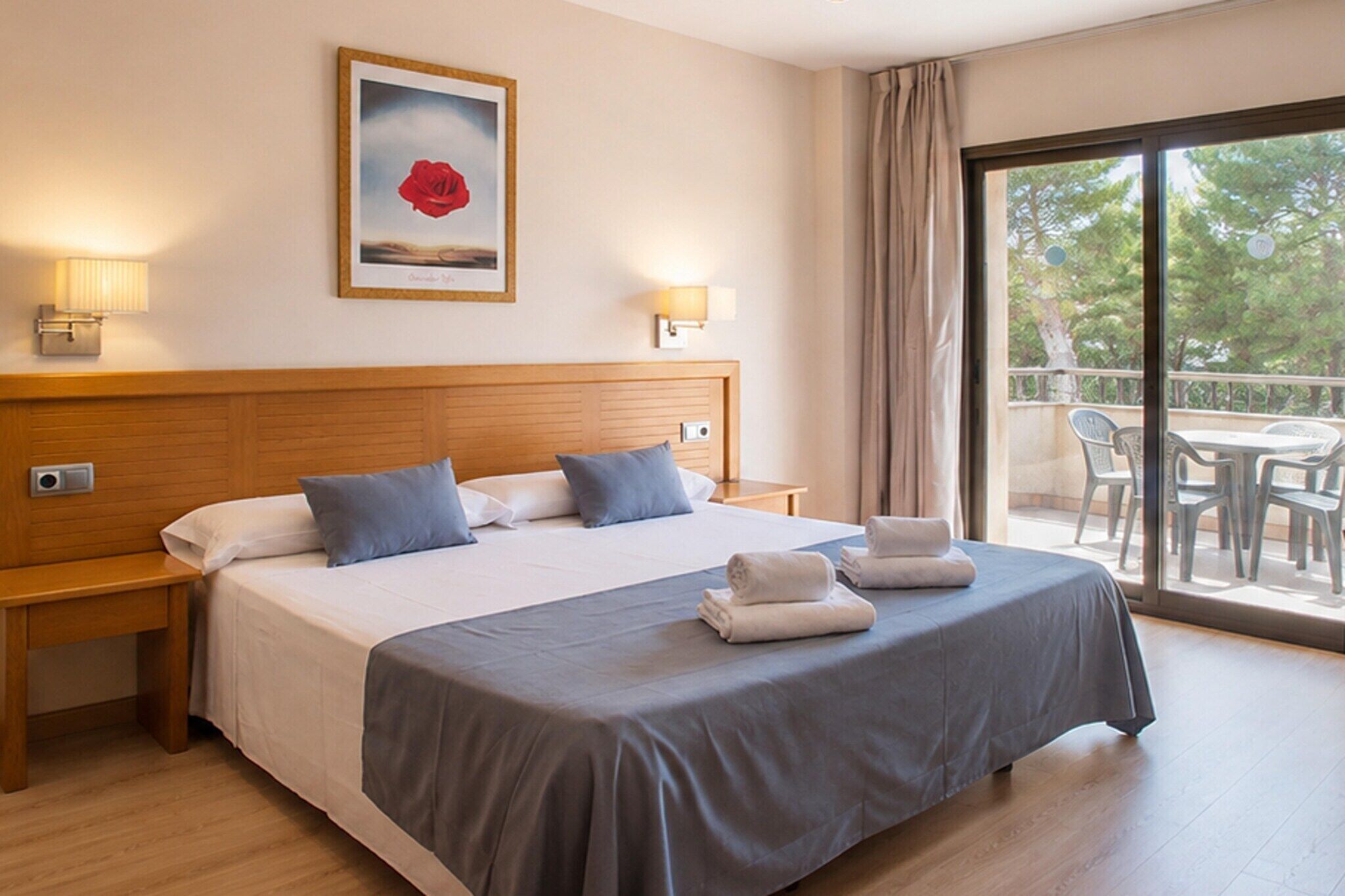 A hotel room with a bed, two bedside tables, a painting, a balcony with chairs, and a view of trees.