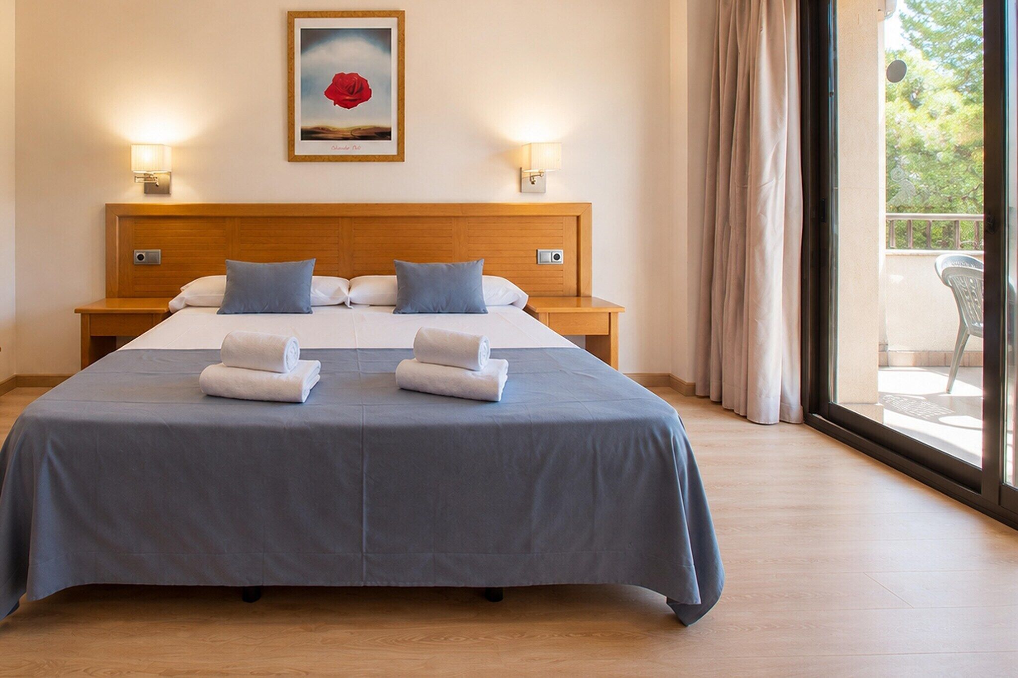 Un cuarto de hotel con una cama grande, dos mesitas de noche, un cuadro en la pared y vistas por la ventana.