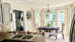 Une cuisine dotée d’un évier à deux bacs, un coin repas avec une table dressée pour deux personnes, et une vue sur l’extérieur grâce à de grandes fenêtres.