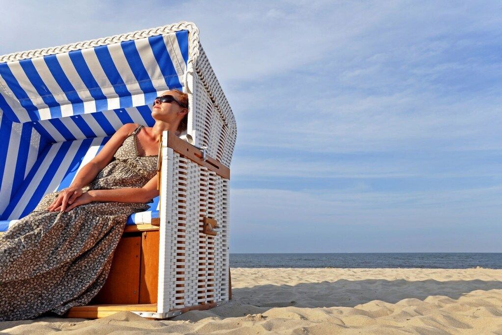 Une chaise de plage dotée d’un auvent à rayures, d’un siège en bois et d’une personne assise dessus.