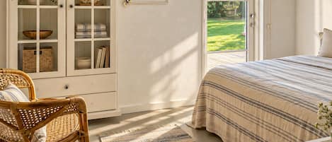 A bedroom with a large window, a wicker chair, a bed with striped bedding, and a cabinet with glass doors.