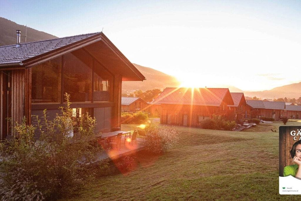 Un bâtiment moderne en bois, doté de grandes fenêtres, d’un espace extérieur couvert avec des sièges et offrant une vue sur un coucher de soleil se déclinant sur un paysage vallonné.