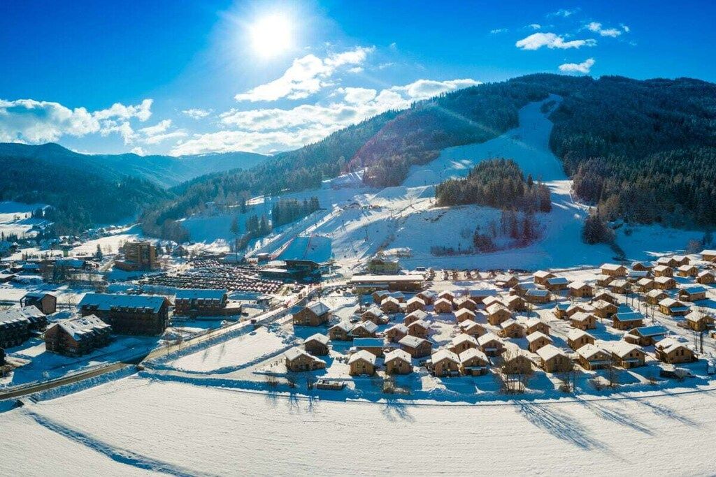 A snow-covered village with traditional wooden houses, a ski slope, and a clear blue sky.