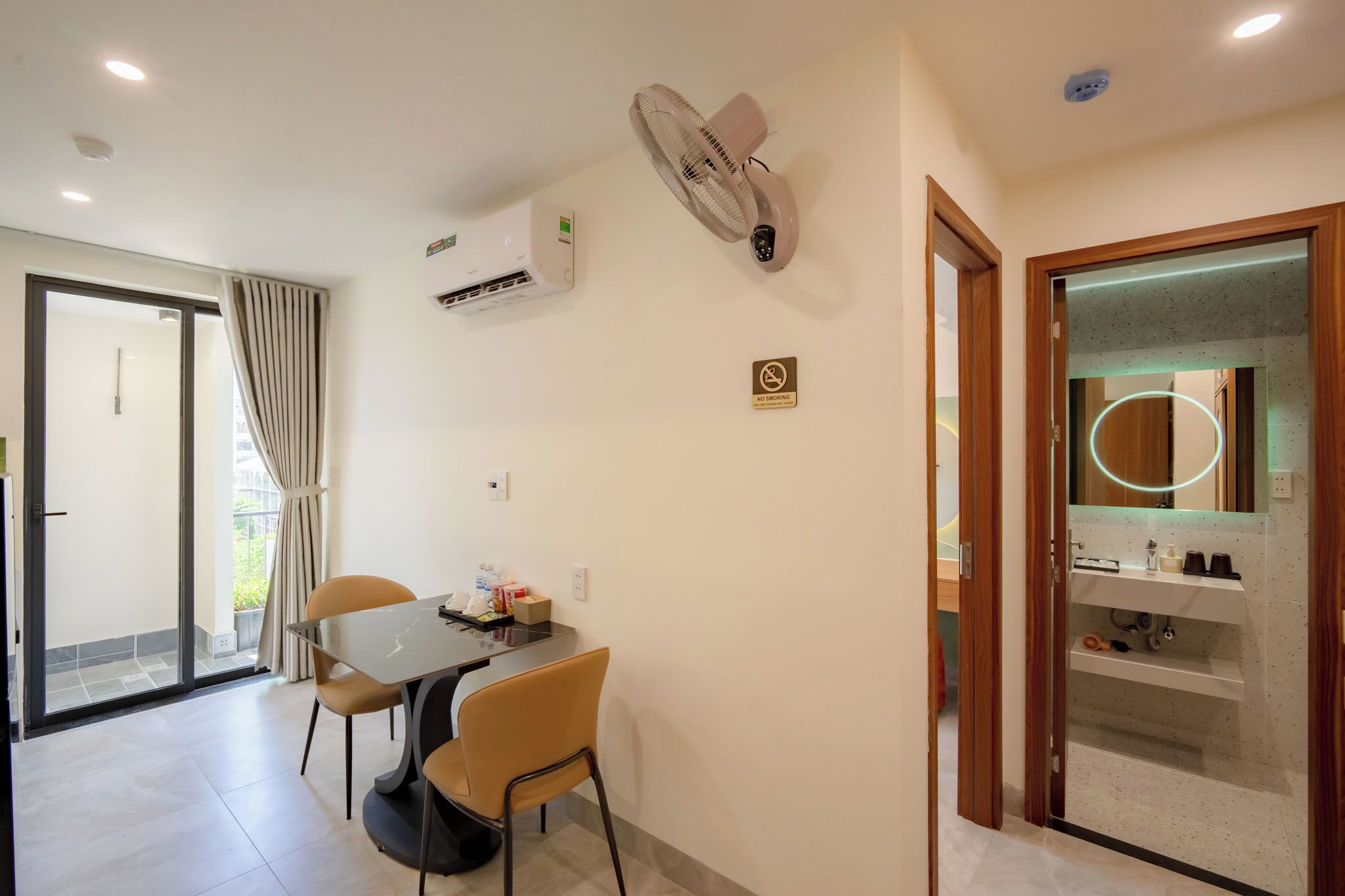 Habitación de hotel con zona de comedor, baño con espejo y artículos de aseo, y puerta corrediza de vidrio al exterior.
