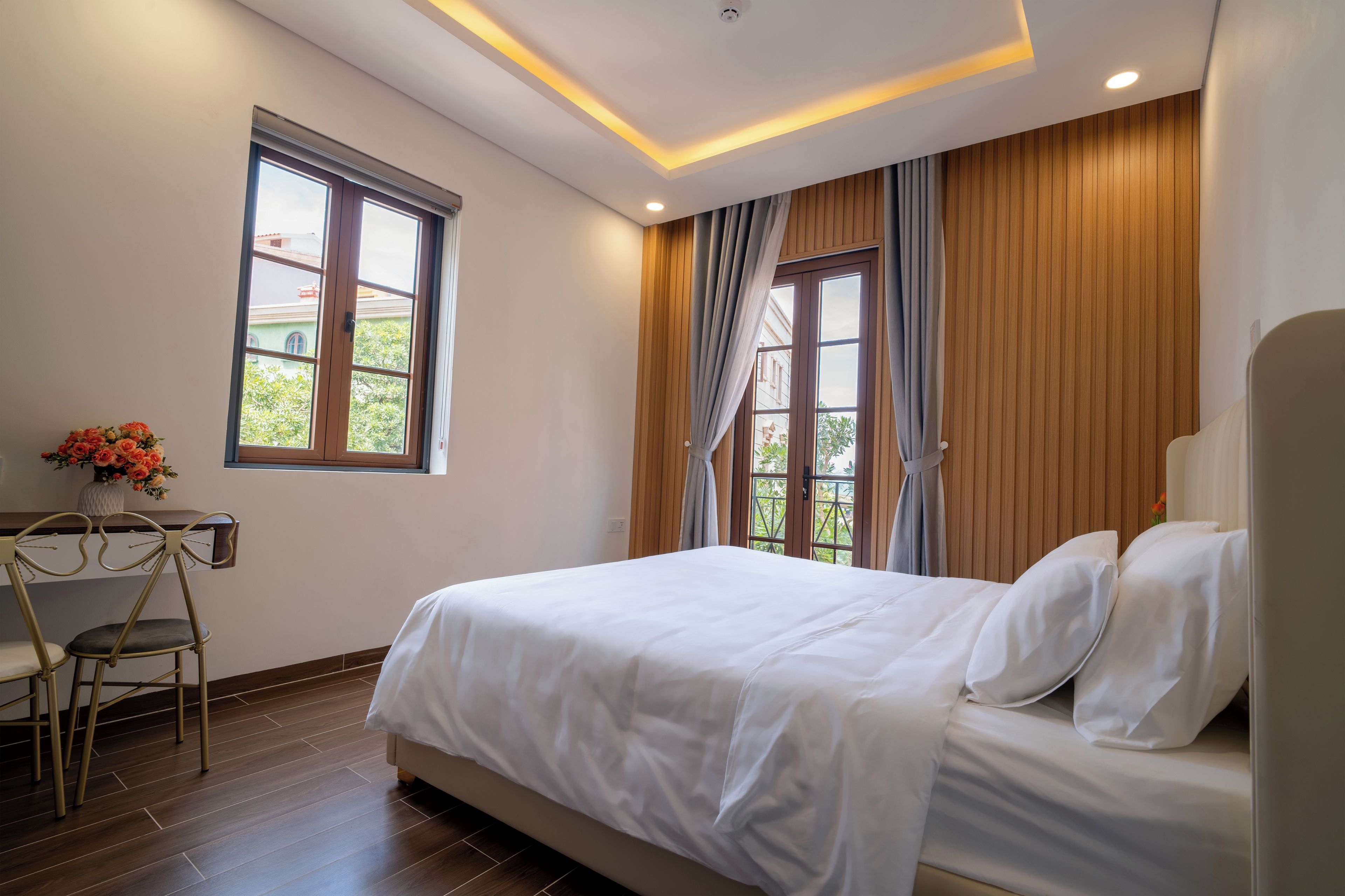 Habitación de hotel con cama, escritorio y silla. Ventanas con cortinas y vistas al exterior con vegetación.